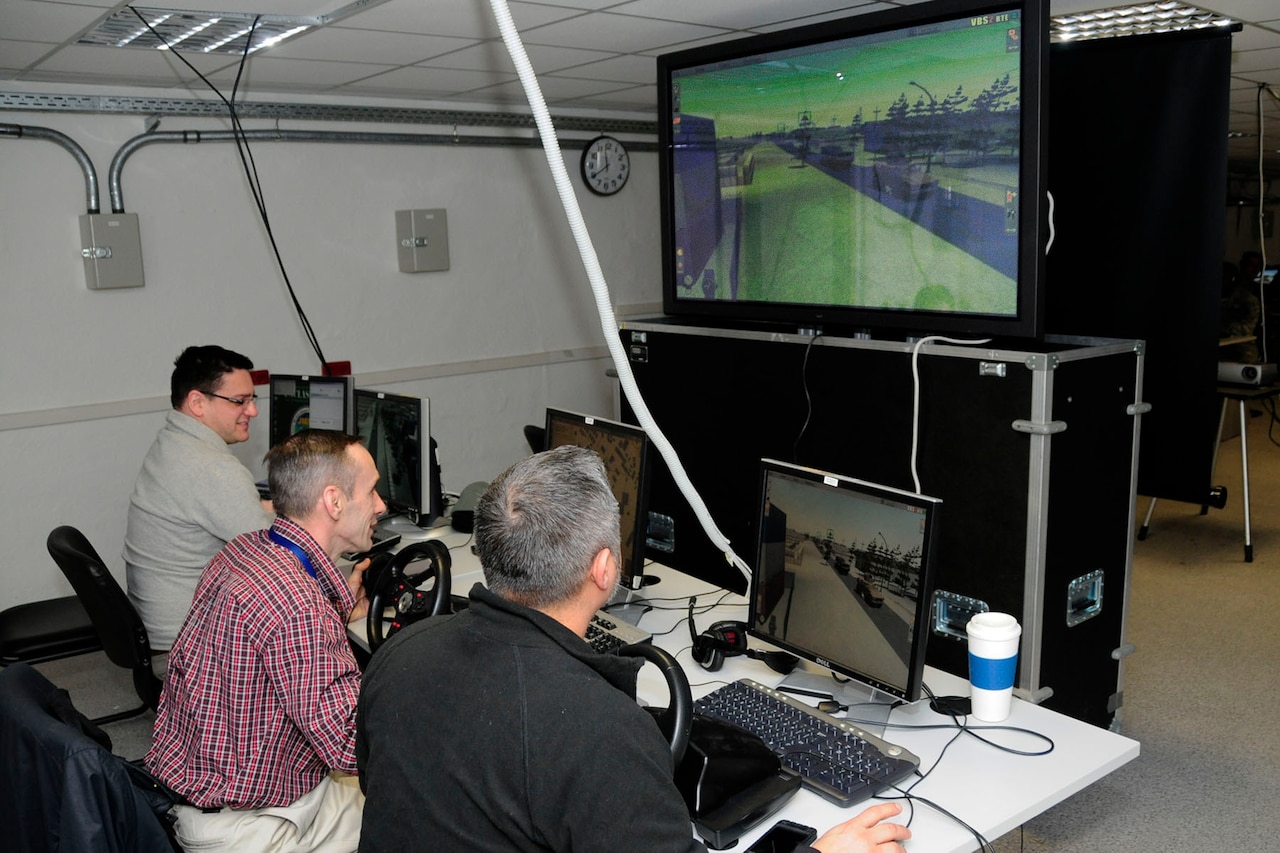 Three men sit in front of computer terminals and a large TV screen.