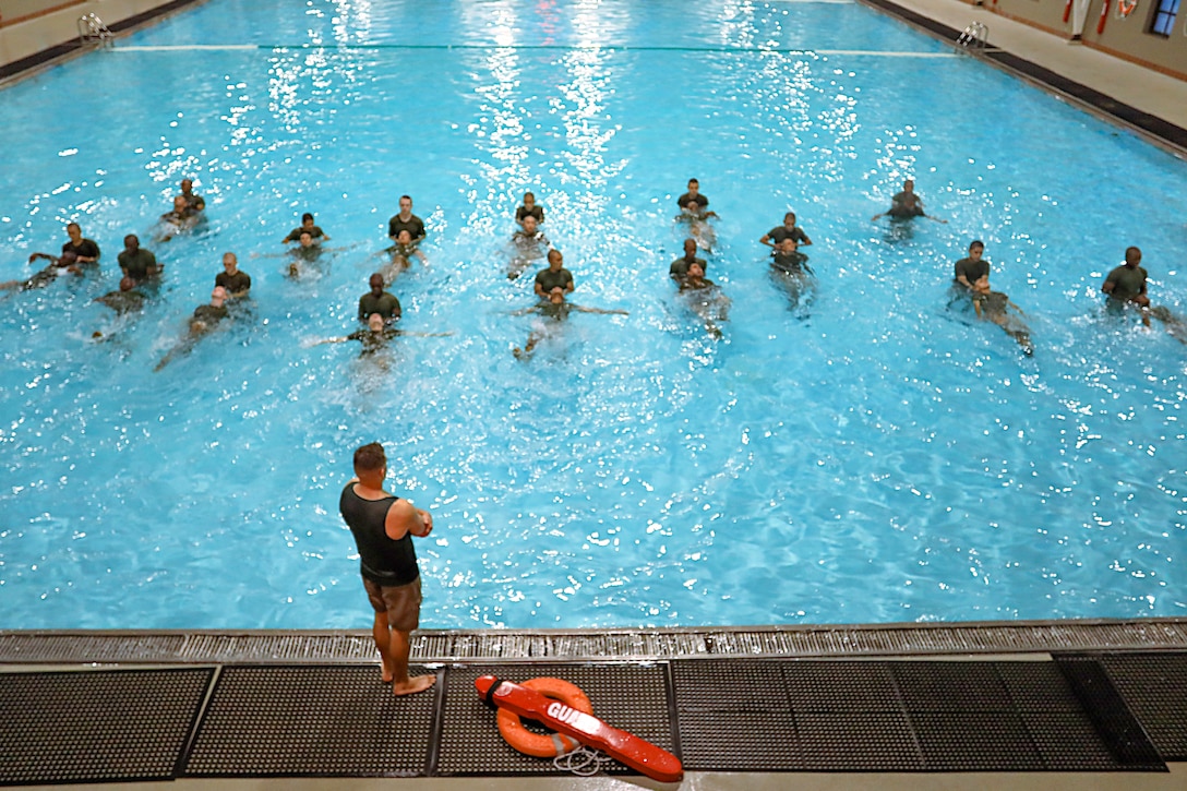 A group of Marines in a swimming pool while an other stands on the edge.