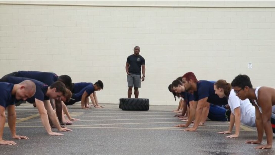 A man standing behind a tire on the ground watches several others around him do pushups.