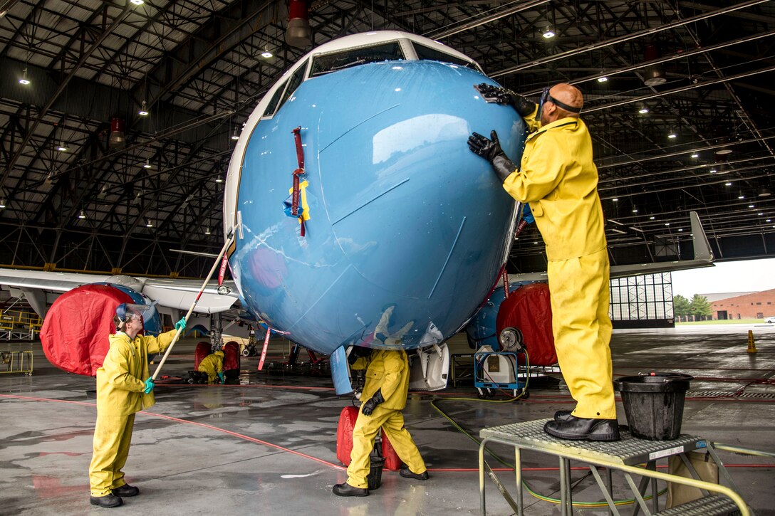 Three airmen in yellow coveralls wash the blue nose of an aircraft in a hangar.
