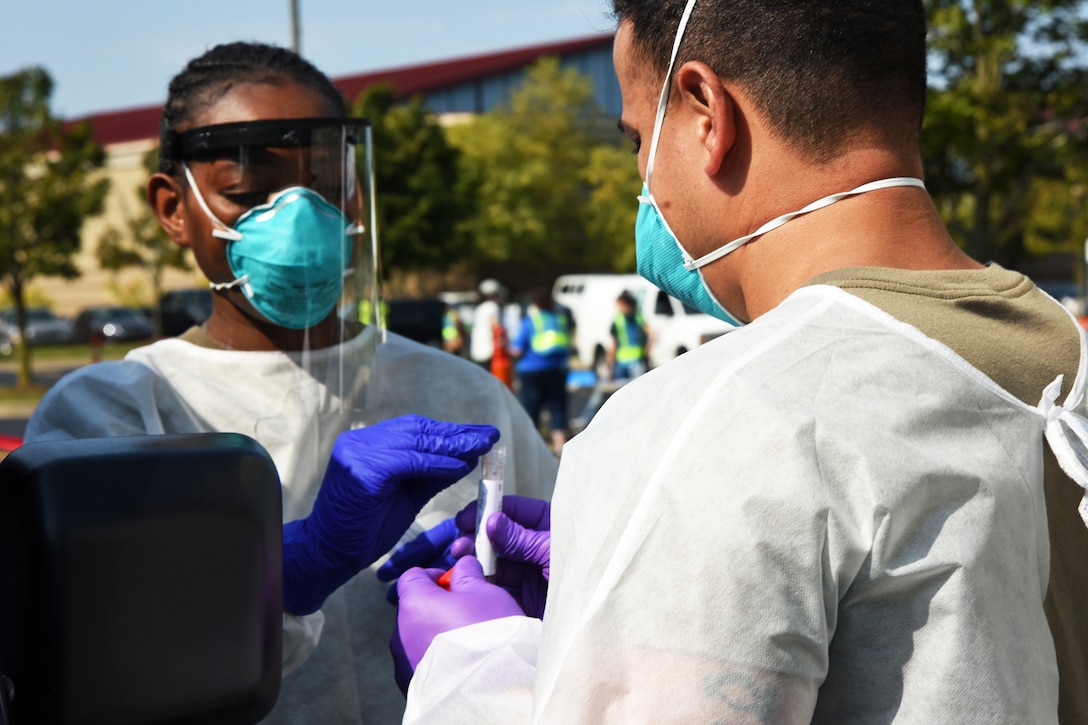 A woman wearing personal protective equipment puts something into a test tube being held by a man, who is also wearing personal protective equipment.