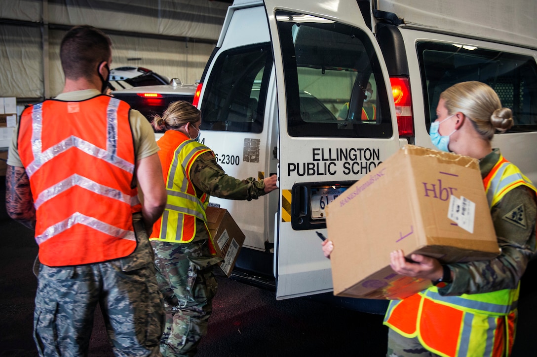 Three service members -- two women and one man --  wearing face masks and reflective vests carry boxes to a vehicle.