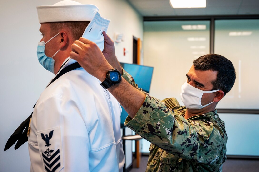 A Navy chief wearing a face mask inspects the uniform of another sailor wearing a face mask.