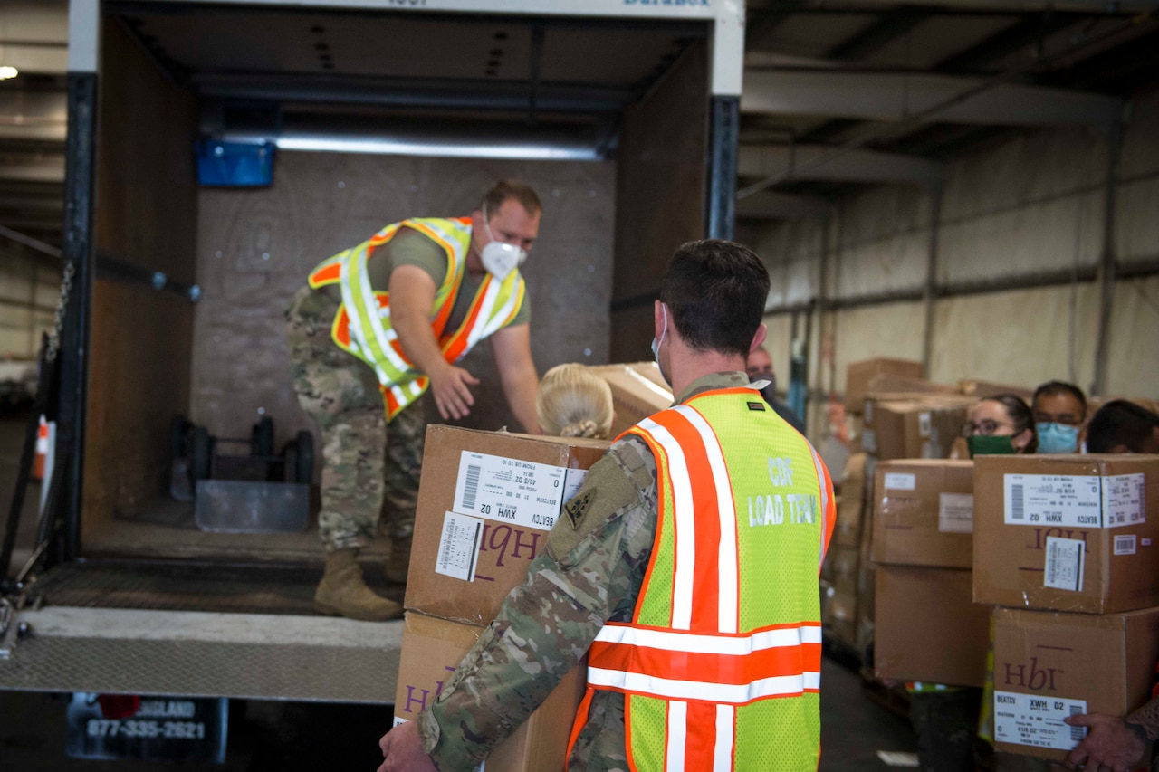 Soldiers and airmen wearing protective equipment load boxes into a vehicle.