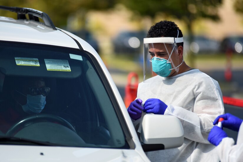 A man wearing personal protective equipment looks into a vehicle's window at a driver wearing a face mask.