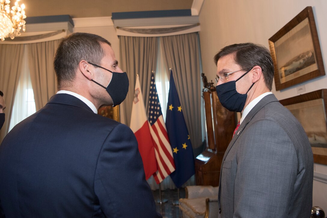 Two men, wearing masks, meet. The flags of Malta, the U.S. and the European Union are in the background.