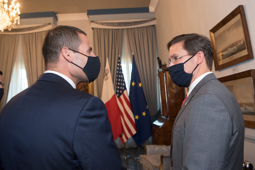 Two men, wearing masks, meet. The flags of Malta, the U.S. and the European Union are in the background.