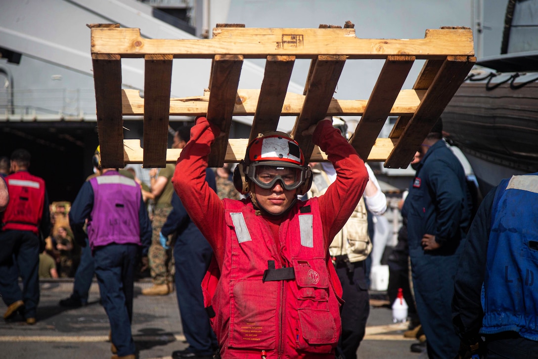 A sailor holds up a wooden pallet while aboard a ship.