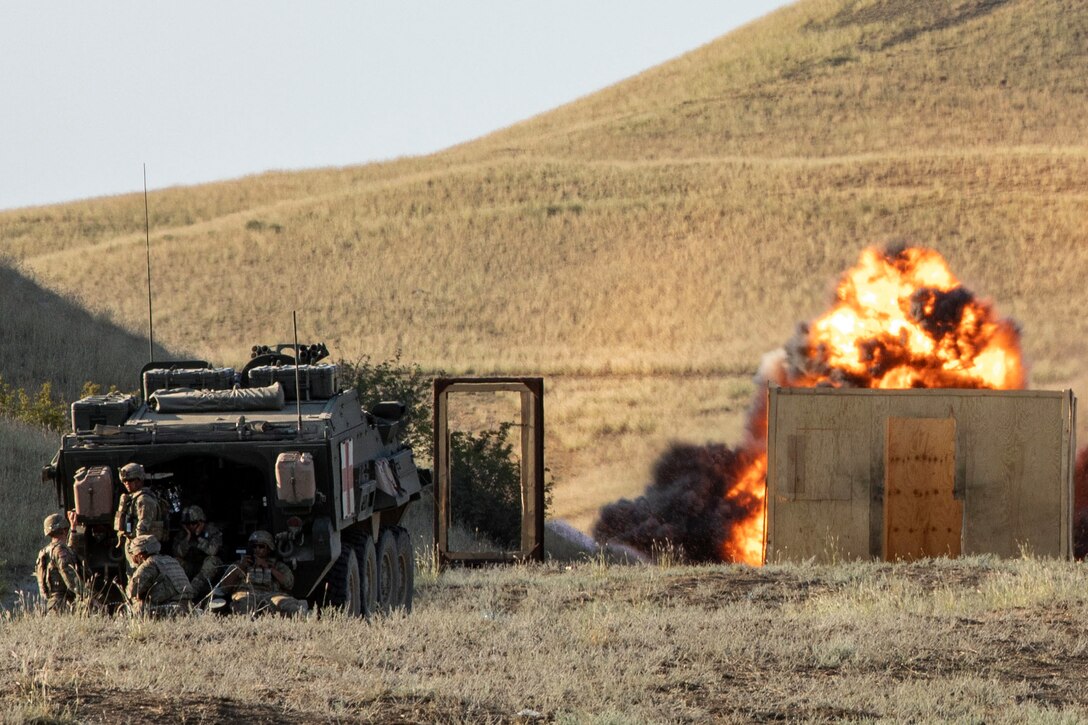 Soldiers take cover behind a tank while an explosion goes off during training.