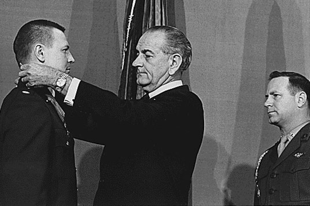 A man puts a medal around another man’s neck as a third man watches.