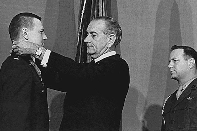 A man puts a medal around another man’s neck as a third man watches.