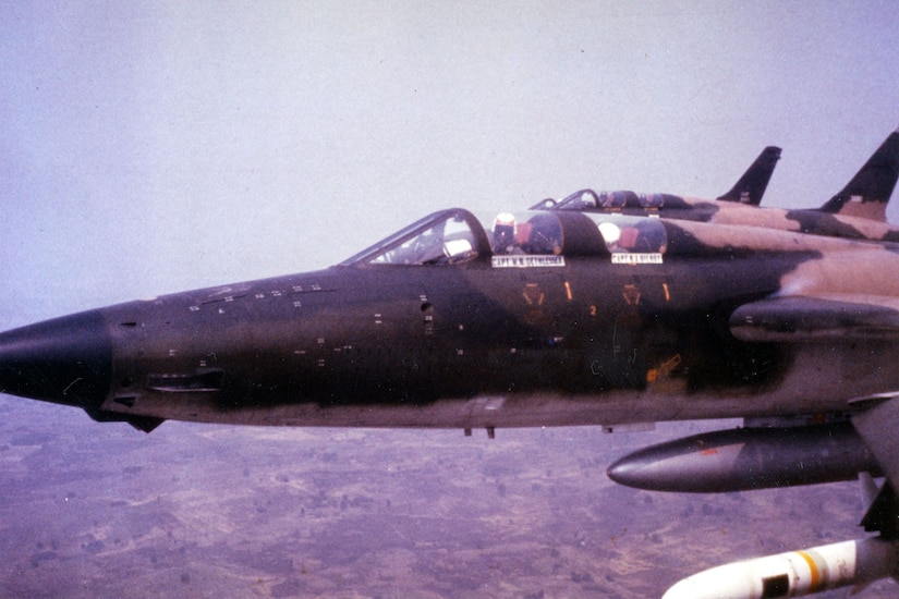 A fighter jet holding two passengers flies over land.