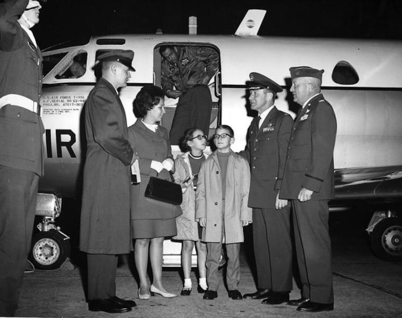 Two military men greet a family of four outside the door of a small airplane.