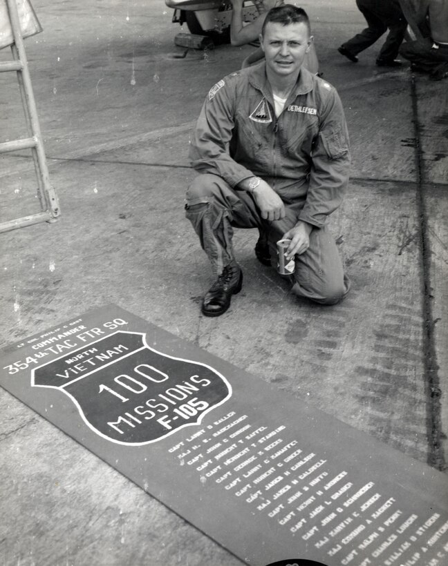 A man kneels on the ground beside a poster noting 100 missions over North Vietnam.