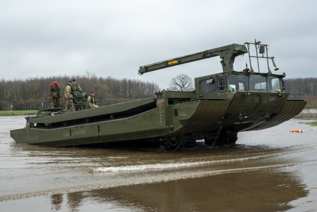 Four people stand atop an amphibious vehicle as it emerges from muddy waters.