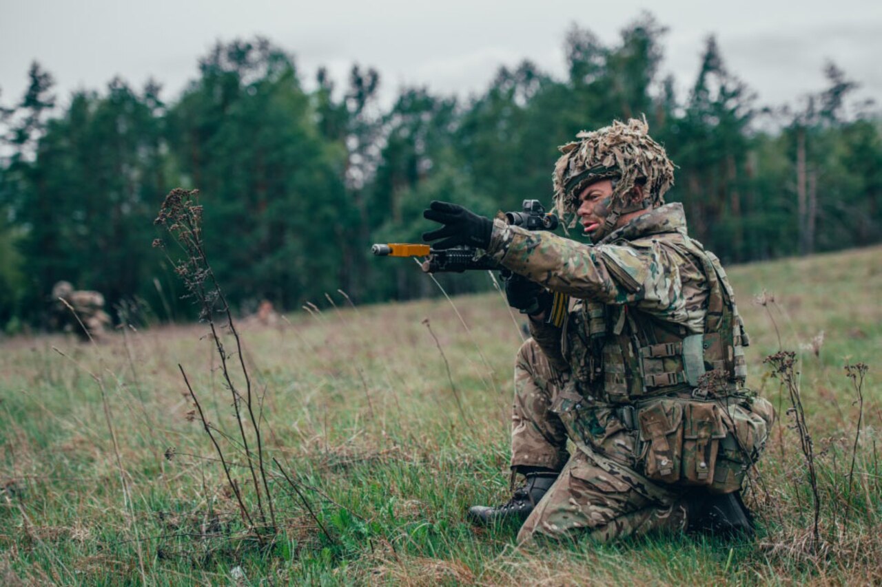 A soldier wearing camouflage gear sits on the ground in a field to aim his weapon.