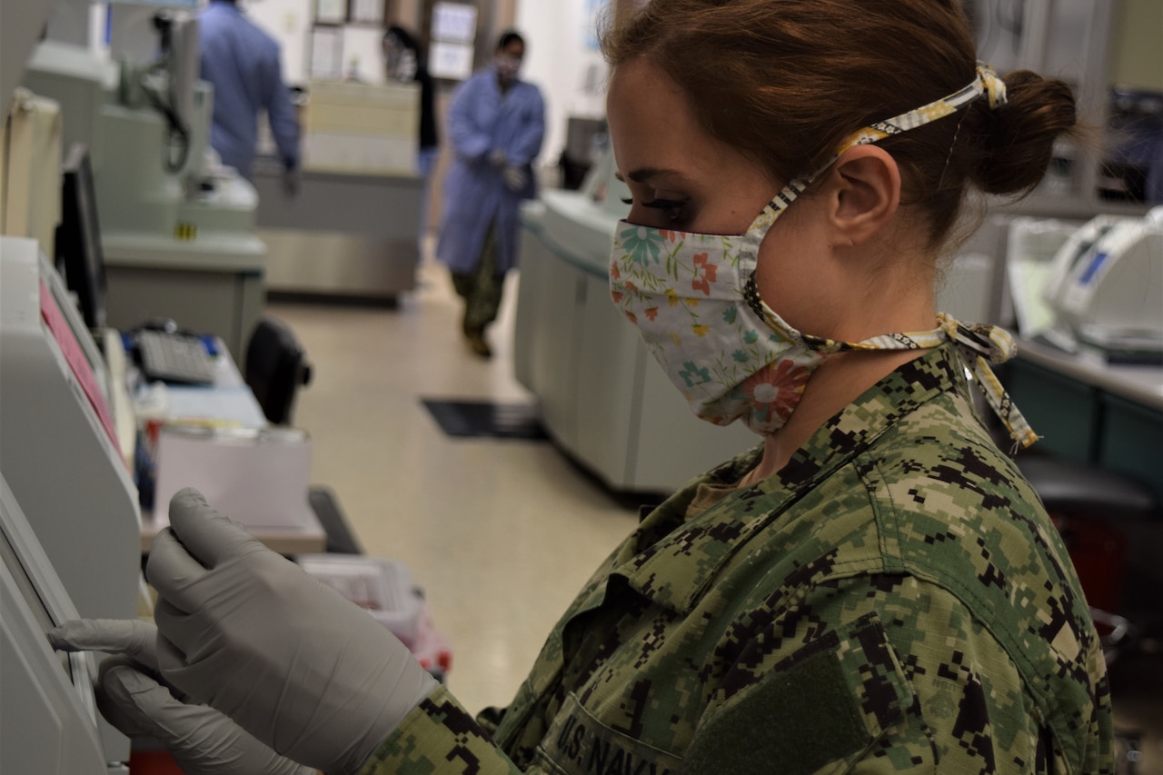 A female sailor wearing a mask checks lab equipment.