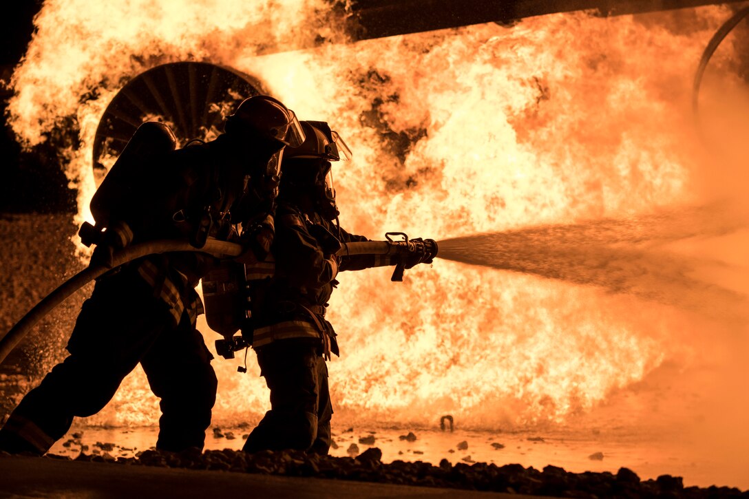 Two Air Force firefighters, shown in silhouette, hold a hose while spraying water on a fire.