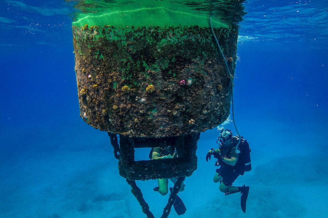 Two Navy divers work in vivid blue water by a large buoy.