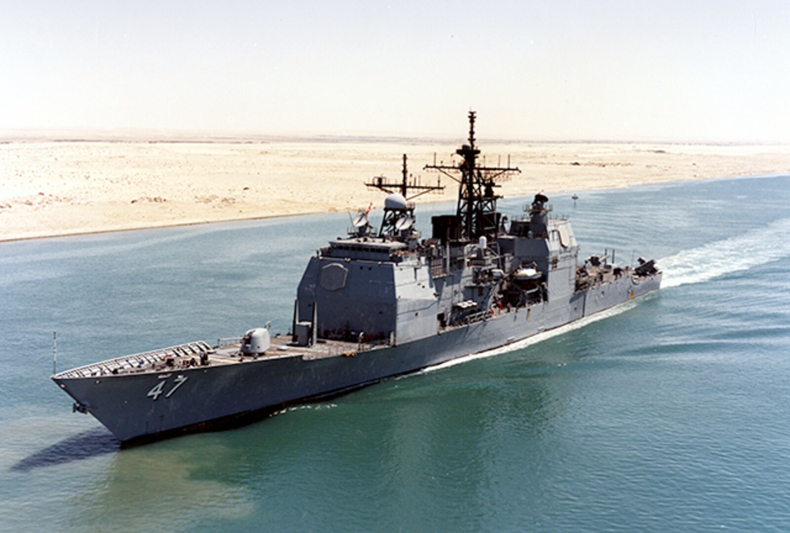 A Navy ship moves from right to left in blue water with sand of the desert behind it.