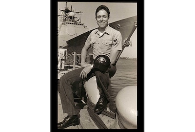 A young sailor in a working uniform sits on the pier with a ship tied up behind him