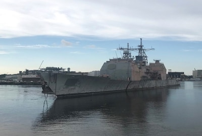 The ex-USS Ticonderoga (CG47) sits in an industrial dock area showing her age.