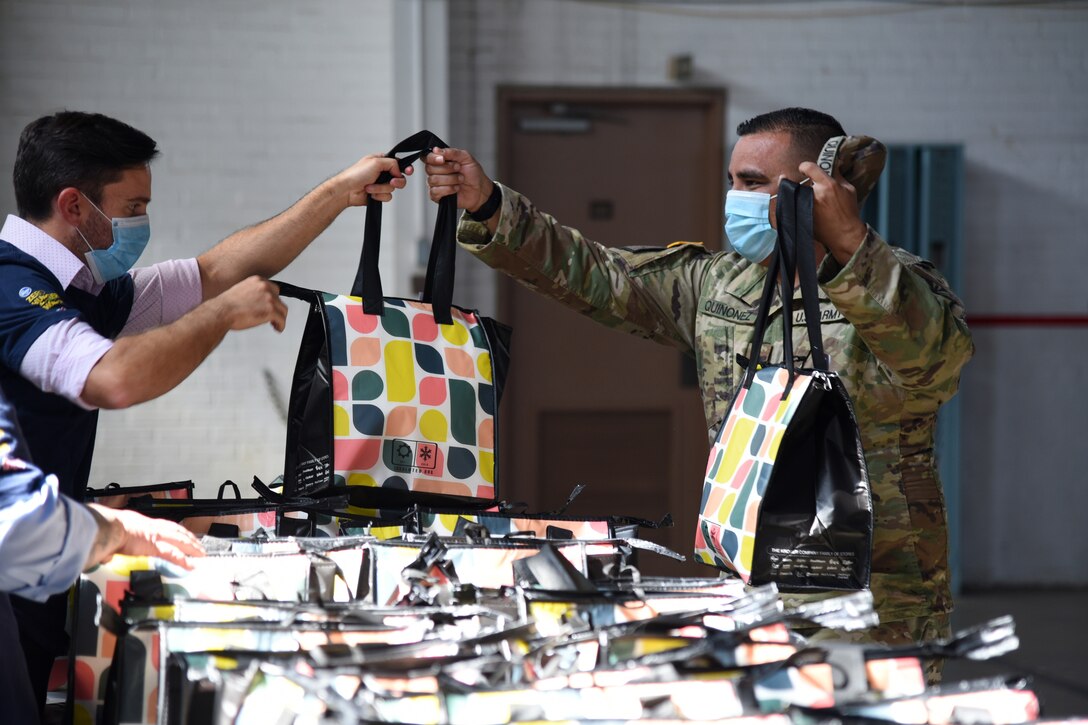 A member of the Arizona USO, wearing a face mask and gloves, gives a colorful bag to a soldier wearing a face mask.
