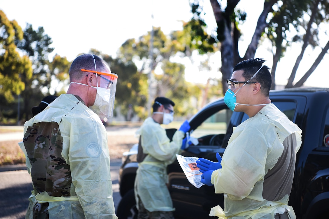 Two men wearing personal protective equipment talk while another man stands holding a test tube at the window of a vehicle.