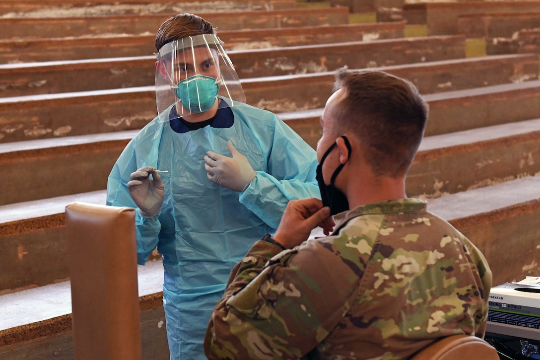 A person wearing personal protective equipment holds a swab while talking to a soldier.