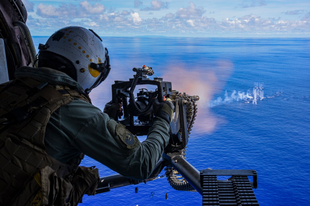 A sailor fires a machine gun from a helicopter into the ocean.