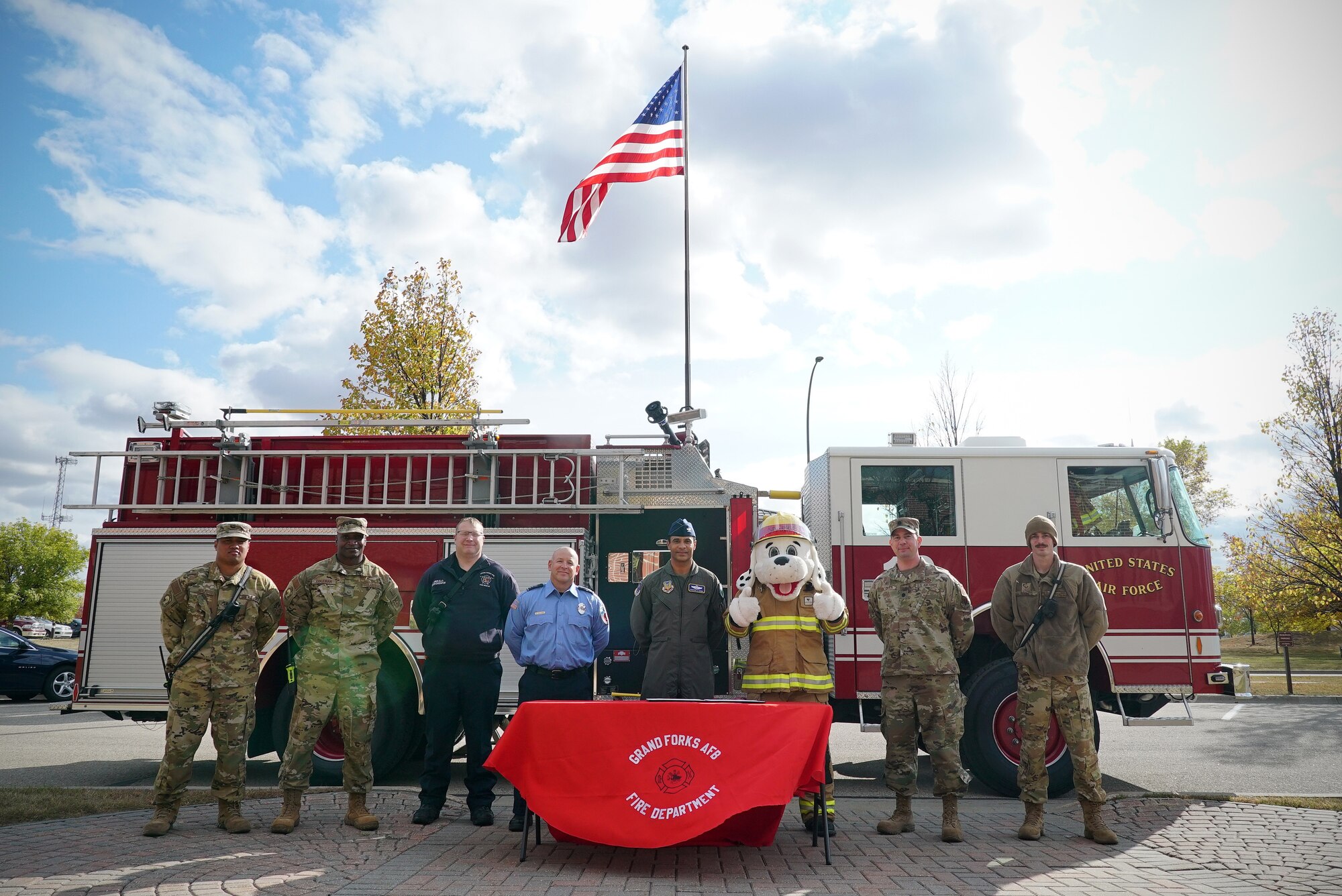 Seven men and one person in a firefighting dog mascot costume stand shoulder-to-shoulder with a red fire engine behind them.