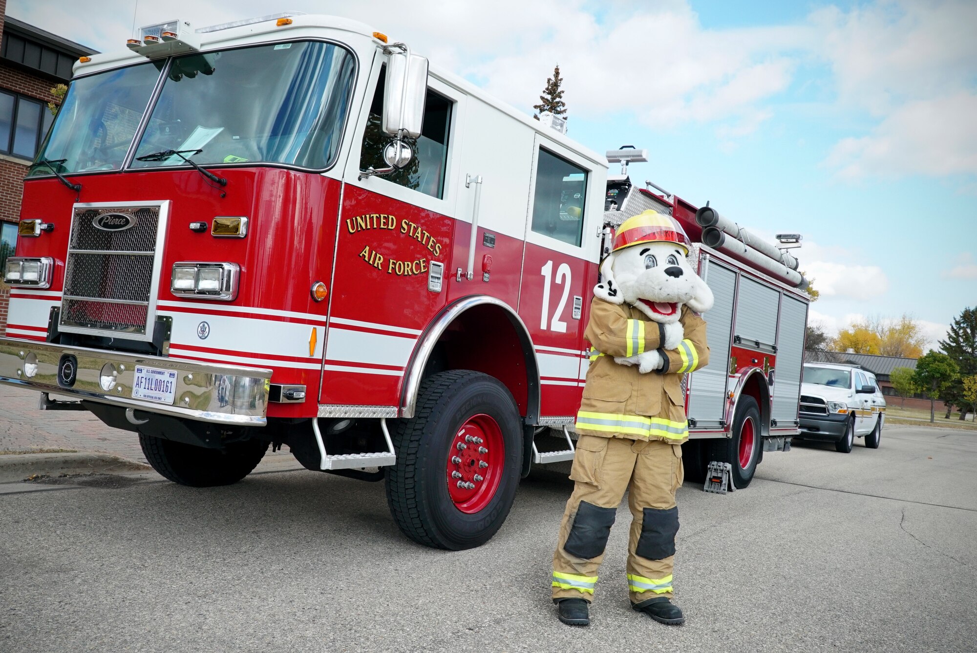 A person in a firefighting dog mascot stands in front of a red fire engine.