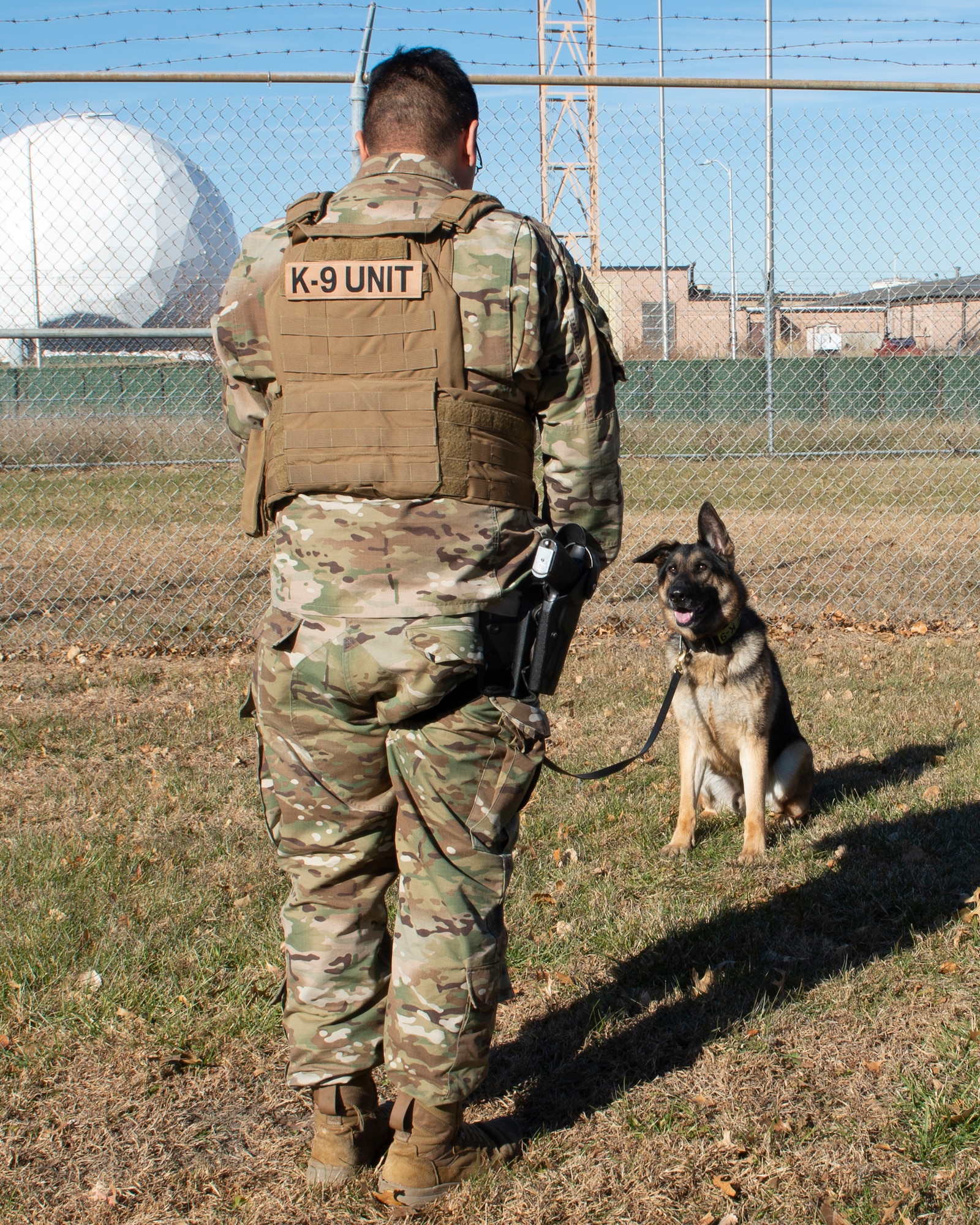 in the foreground a Airman wearing a vest with K9 unit on the backside of vest, he standing in front of a military working dog sitting in front of the Airman
