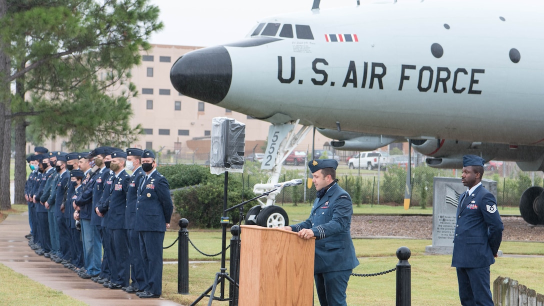 Man talking at podium with aircraft behind him and a line of people next to him.