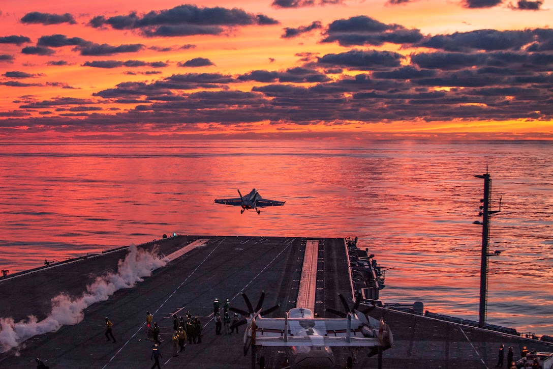 An aircraft takes off from a ship's flight deck toward a pink- and orange-streaked sky.
