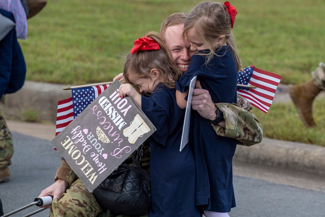 An airman kneels on pavement outside and smiles while embracing two girls in matching blue dresses and red bows.
