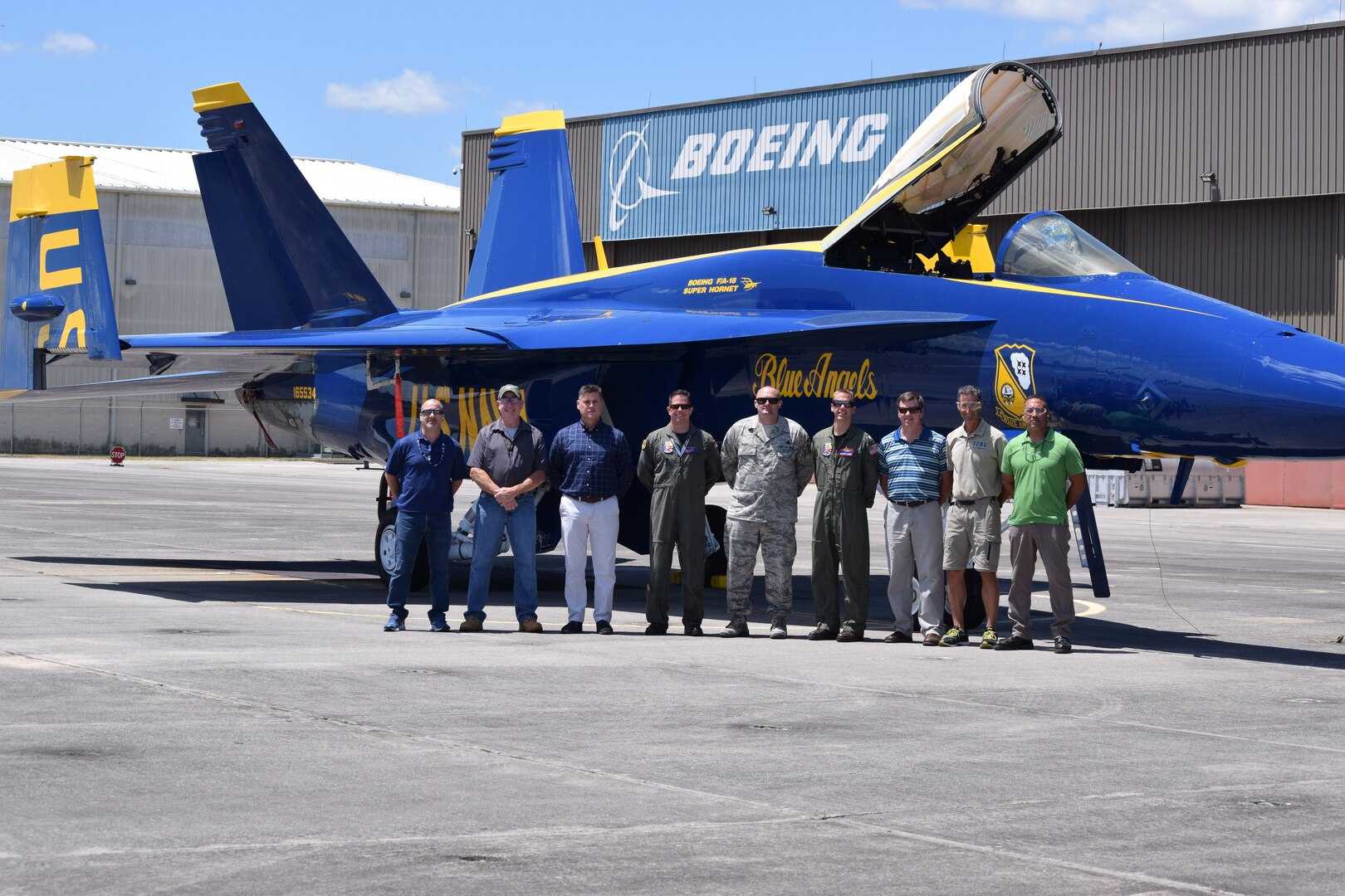 Nine people stand in front of a blue fighter jet with yellow trim