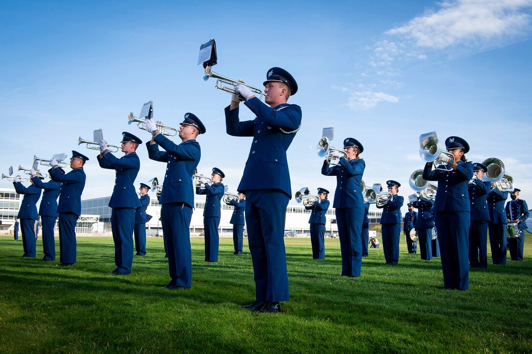 A large group of airmen play instruments as they stand in formation on a  large field.