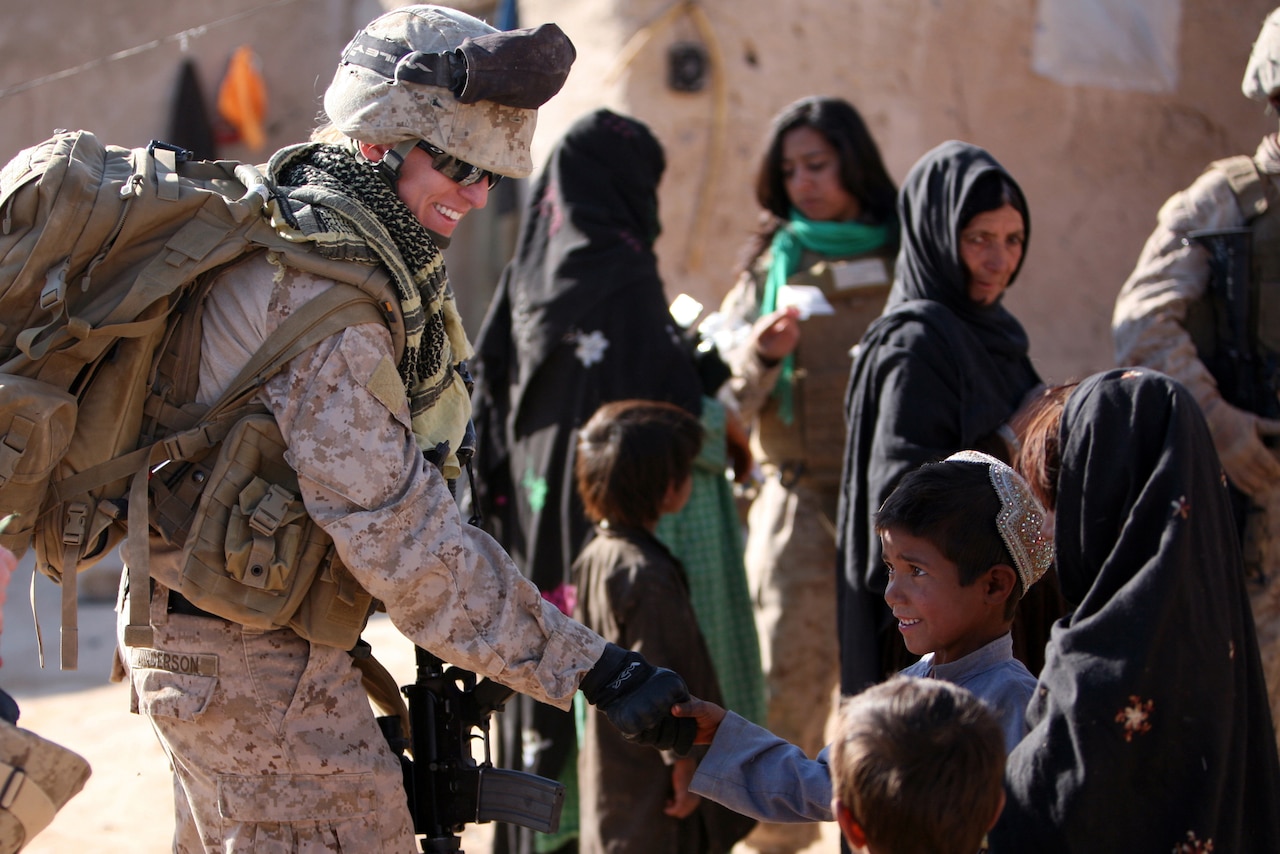 A woman in a military uniform shakes hands with a young boy. Other women and children are milling about in the background.