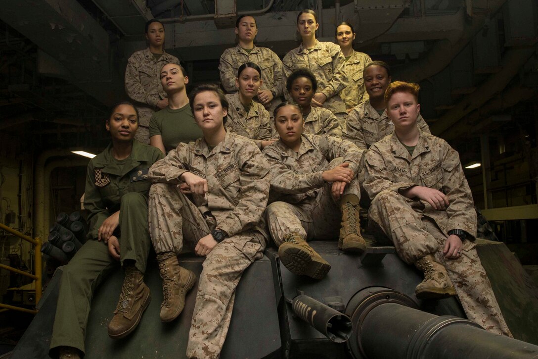 A dozen women in military uniforms sit on a tank turret.
