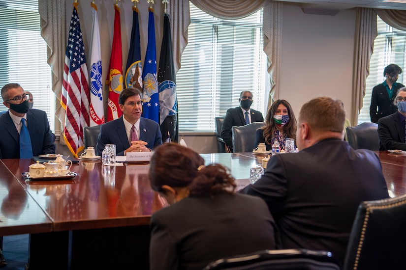 People wearing face masks sit at a conference table while one without a mask speaks to a man just across the table from him.