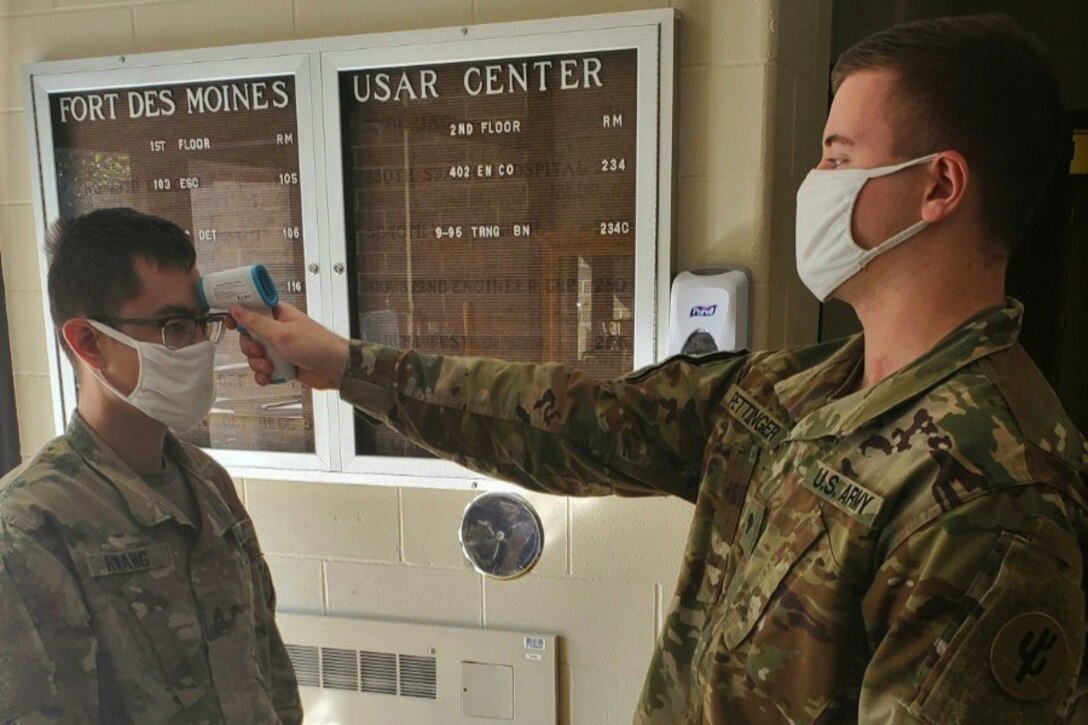 A soldier wearing a face mask takes the temperature of another soldier wearing a face mask.