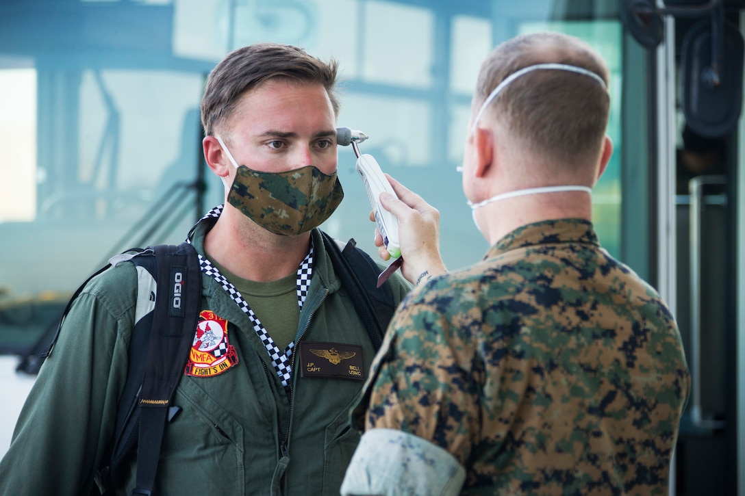 A  Marine holds a digital thermometer to the temple of a sailor; both men are wearing face masks.