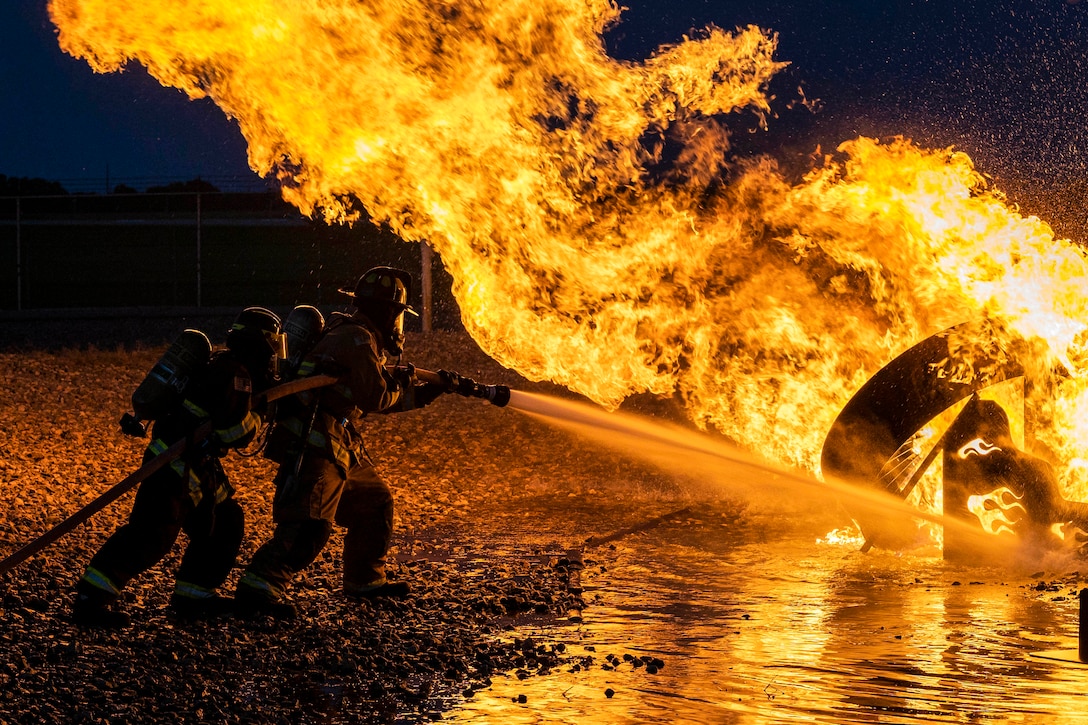 Two firefighters train a water hose on a large fire lighting up a dark blue night sky.