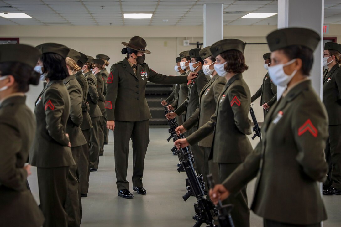 Marines stand in rows in dress uniforms as a drill sergeant inspects their uniforms.
