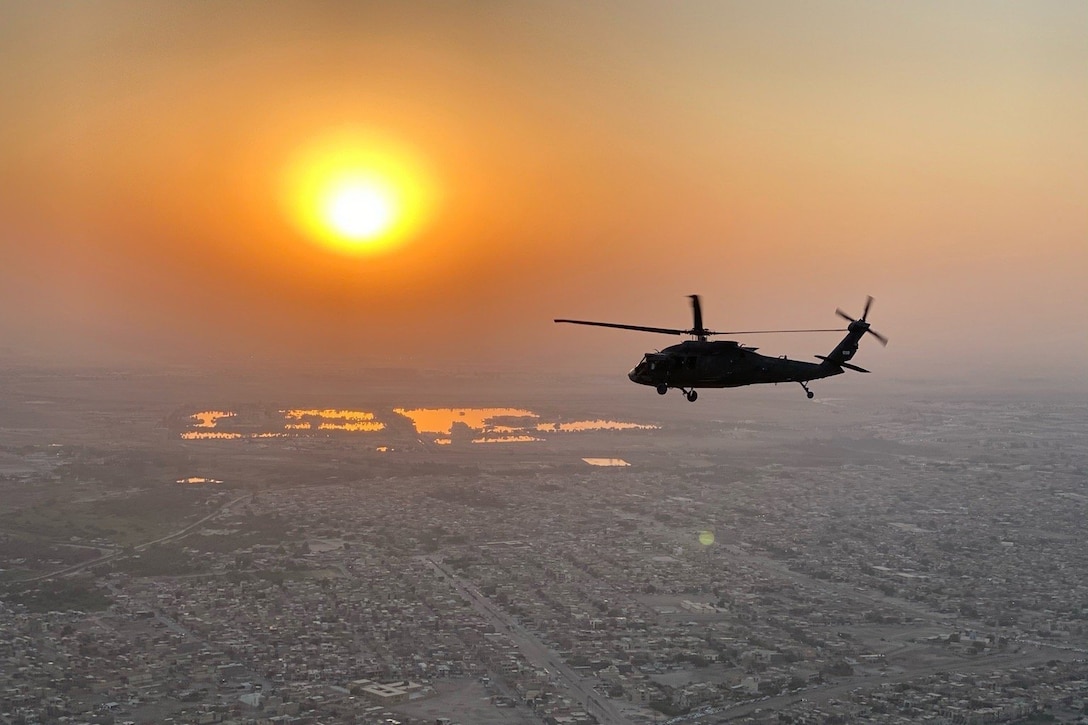 A  military helicopter flies above a city with the sun in the background.