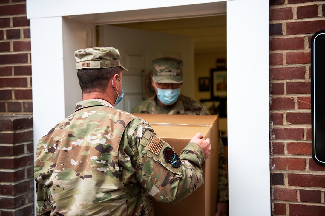 Two male airmen wearing face masks carry a large box through a doorway.