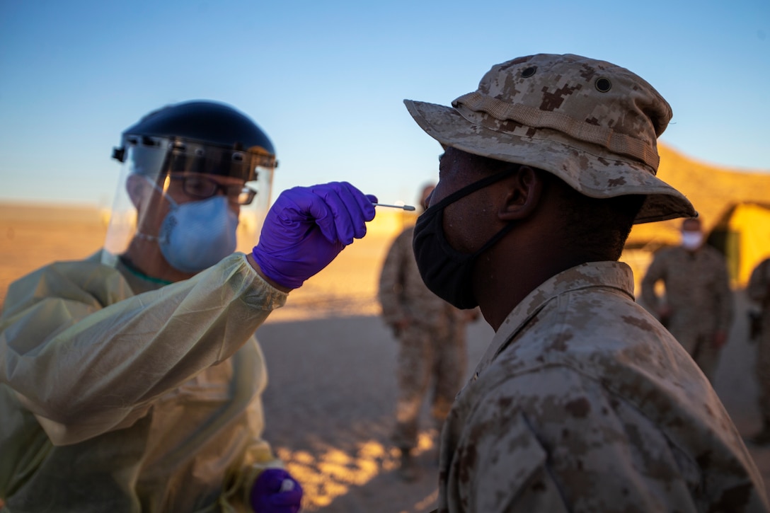 A Marine wearing personal protective equipment prepares to swab the nose of another Marine, who is also wearing a face mask.