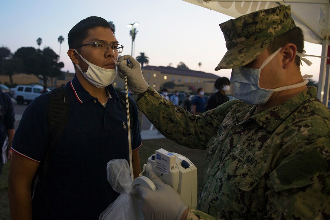 A male Marine wearing a face mask and gloves takes the temperature of a new, male Marine recruit.