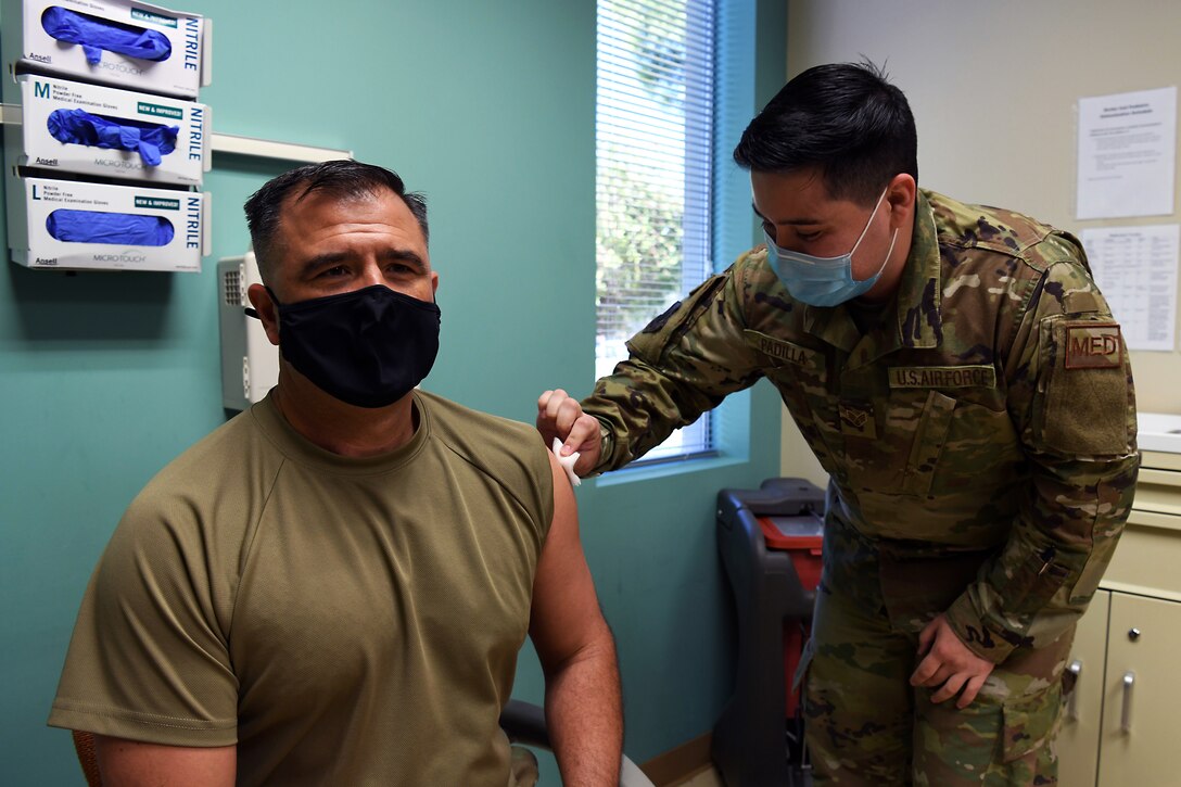 A male airman wearing a face mask swabs the upper arm of another male service member.